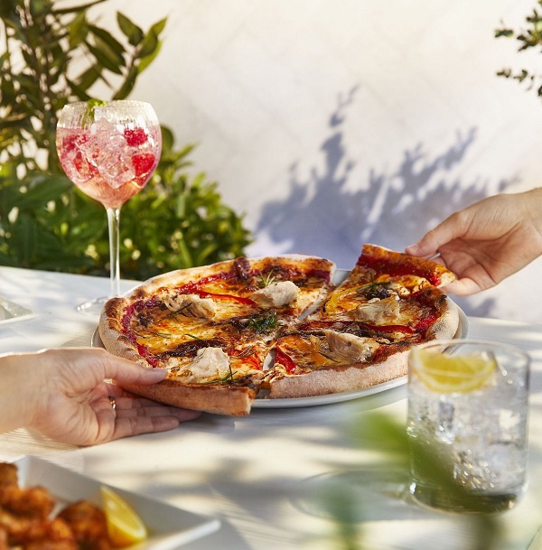 two hands taking a slice of pizza each from a plate and drink with fruit and ice in a gin glass on the table.