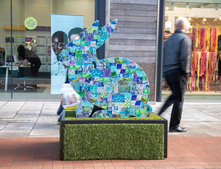 The whiteley rabbit sculpture- a rabbit shaped sculpture in situ at the whiteley centre. It has patches of different blue colour on it and is on a green plinth.